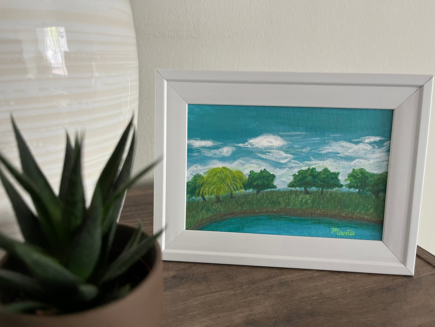 Framed pond painting in display next to vase and succulent plant
